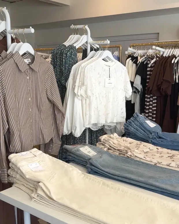 A display table stacked with folded jeans in cream and blue washes, in front of rails of striped shirts and floral dresses.