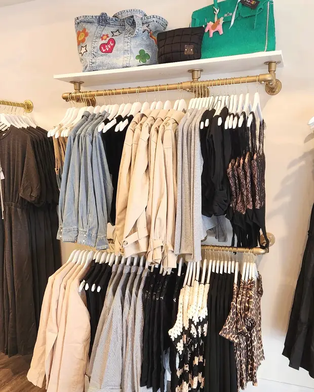 Brass clothing rails inside the boutique, hung with denim jackets, trench coats, knits and lace tops, with handbags on the white shelf above.