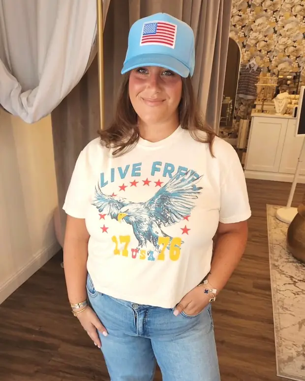 A woman in the shop wearing a cream graphic tee and light-wash jeans, standing in front of the flower wall.