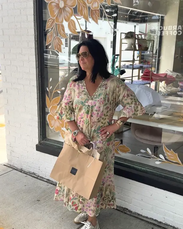 A customer outside the boutique window in a floral kaftan, holding a kraft-paper shopping bag, with the painted gold flowers of the shop window behind her.
