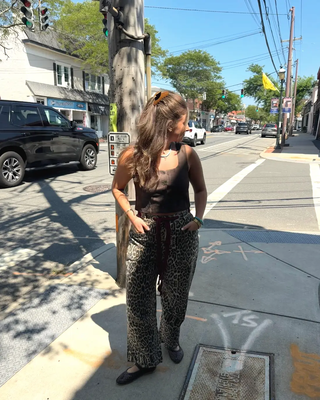 A customer stands on the sidewalk outside the shop in a black top and leopard-print wide-leg trousers, with shops and traffic on the street behind her.